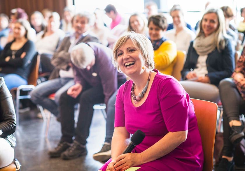 Babara Guwak sitzt lachend im Publikum und h&auml;lt dabei ein Mikrofon in der Hand